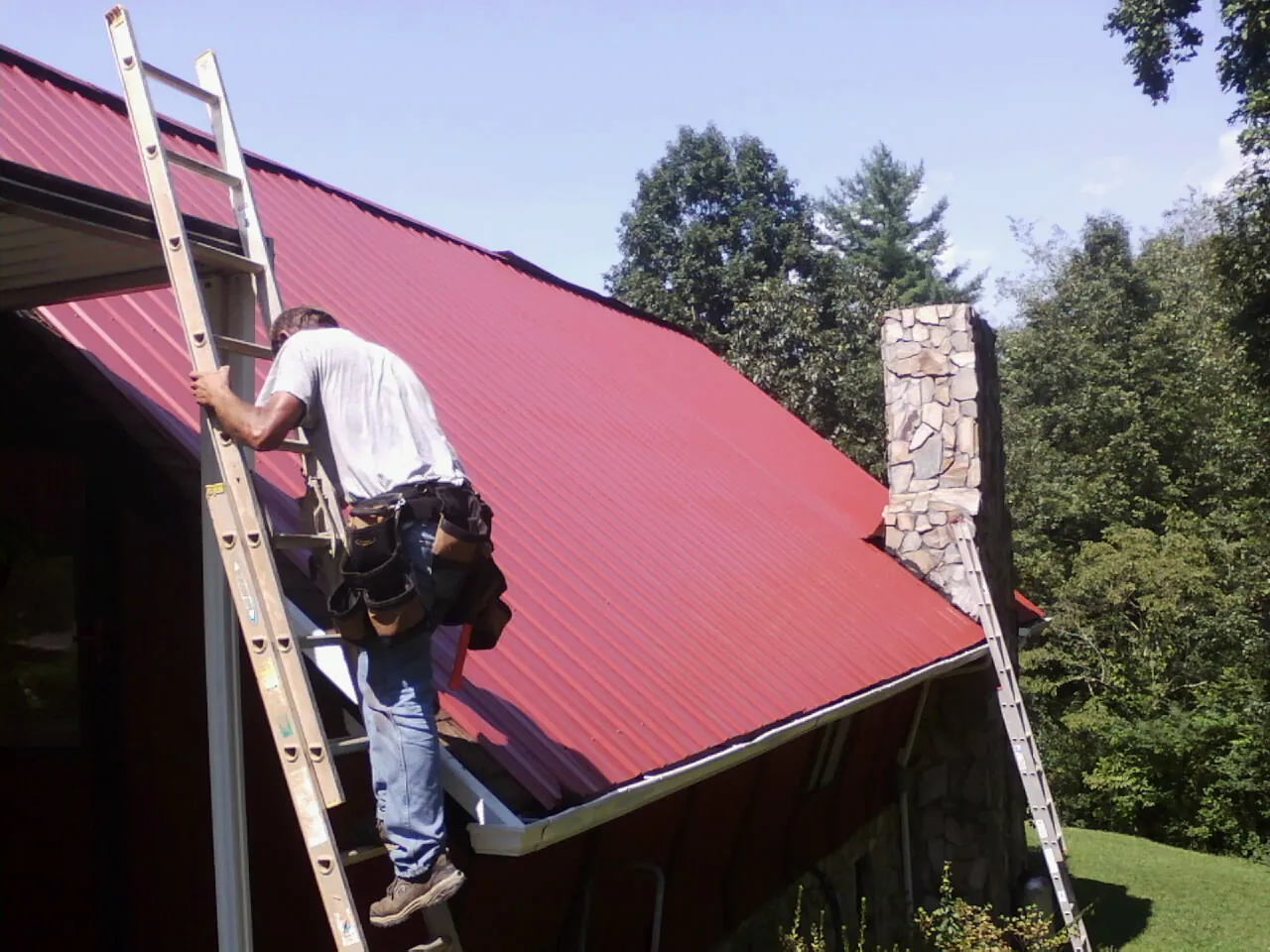 Roofer on a ladder beside a red metal roof and stone chimney — Soffit & Fascia Repair by Roofing Brothers in Bellefonte